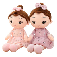 Two plush toys with brown hair and pink dresses on a wooden surface.